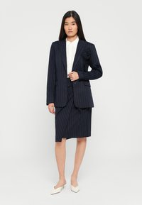 Kvinne i marineblå pinstripe blazer med blomsterdetalj, matchende skjørt, hvit skjorte med knapper og hvite lave hæler, står mot en hvit bakgrunn.