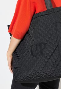 Person trägt einen hellorangefarbenen kurzärmligen Pullover und hält eine große schwarze gesteppte Tasche mit der Aufschrift "UP" an der Seite.