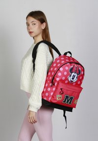 Mochila roja con lunares que presenta un gráfico de Minnie Mouse, detalles negros, un bolsillo frontal con un parche que dice "Clase del 28" y correas ajustables.