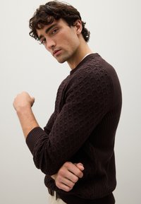 Maglione a maglia marrone scuro con motivo testurizzato, orlo e polsini a coste, design aderente e scollatura rotonda. Il modello è in piedi con le braccia incrociate.