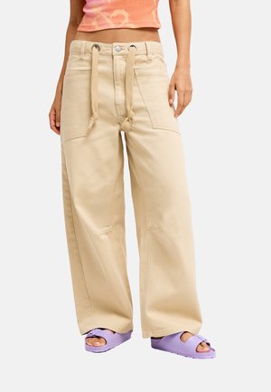 Frau trägt beige, weit geschnittene Hosen mit einem Seilgürtel und lila Sandalen, kombiniert mit einem kurzen orangefarbenen Blumentop.