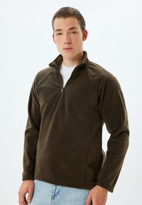 Sudadera de forro polar de un verde oscuro con cuello de media cremallera, mangas raglán y una textura suave, combinada con pantalones de mezclilla azul claro.