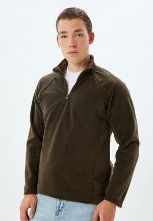 Sudadera de forro polar de un verde oscuro con cuello de media cremallera, mangas raglán y una textura suave, combinada con pantalones de mezclilla azul claro.