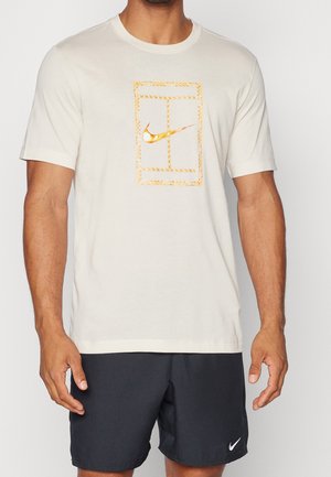 Man draagt een offwhite T-shirt met ronde hals met ontwerp van een gouden tennisbaan en Nike-logo, gecombineerd met zwarte Nike-shorts.