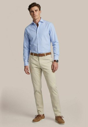 Man wearing light blue striped dress shirt, beige pants, brown belt, brown boat shoes, and wristwatch, standing against plain background.