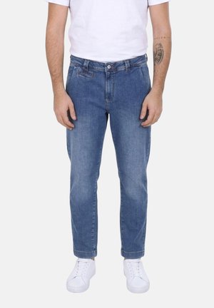 Uomo con jeans blu, sneakers bianche e t-shirt bianca, in piedi con le mani rilassate lungo i fianchi su uno sfondo semplice.