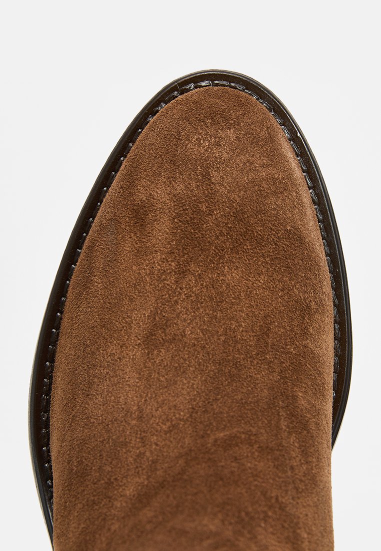Botte en daim marron avec un bout rond, texture lisse et couture noire autour de la semelle. La surface supérieure est douce et mate.