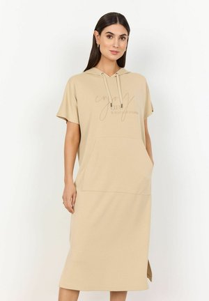 Abito beige con cappuccio e maniche corte, dotato di tasca frontale e spacco laterale. Testo stampato sul petto in un colore a contrasto. Materiale morbido.