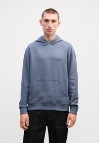 Mīksts zils kapuce krekls, ar priekšējo kabatu un reljefu "Calvin Klein" logotipu. Ribotās piedurknes un apakšmala, brīva piegriezuma.