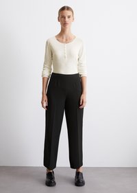 Lys beige langermet topp med Henley-hals og knappdetaljer, paret med høy midje og rette svarte bukser. Svarte slip-on sko.