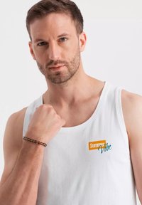 Bílý bavlněný tílko s potiskem "Summer Vibe" v oranžové a tyrkysové barvě. Model má na zápěstí náramek z korálků.