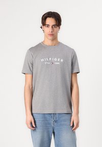 Siva pamučna majica kratkih rukava. Sadrži tekst "HILFIGER USA 1985" u bijeloj boji. Uparen s svijetlo plavim trapericama. Jednostavan, ležeran dizajn.
