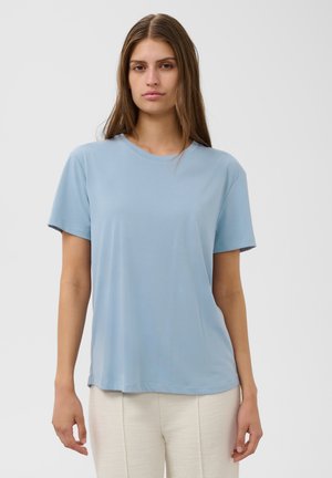 Joven mujer con pelo largo y marrón, vistiendo una camiseta de manga corta de color azul claro y pantalones color crema, de pie frente a un fondo simple.