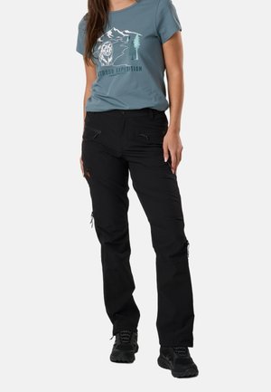 Frau trägt ein blaues Grafik-T-Shirt mit der Aufschrift „Outdoor Expedition“, schwarze Wanderhose mit Reißverschlusstaschen und schwarze Trailschuhe, steht vor weißem Hintergrund.