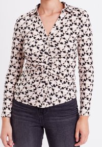 Mujer vistiendo una camisa de botones de manga larga con un patrón geométrico en beige y negro, y jeans oscuros ajustados.