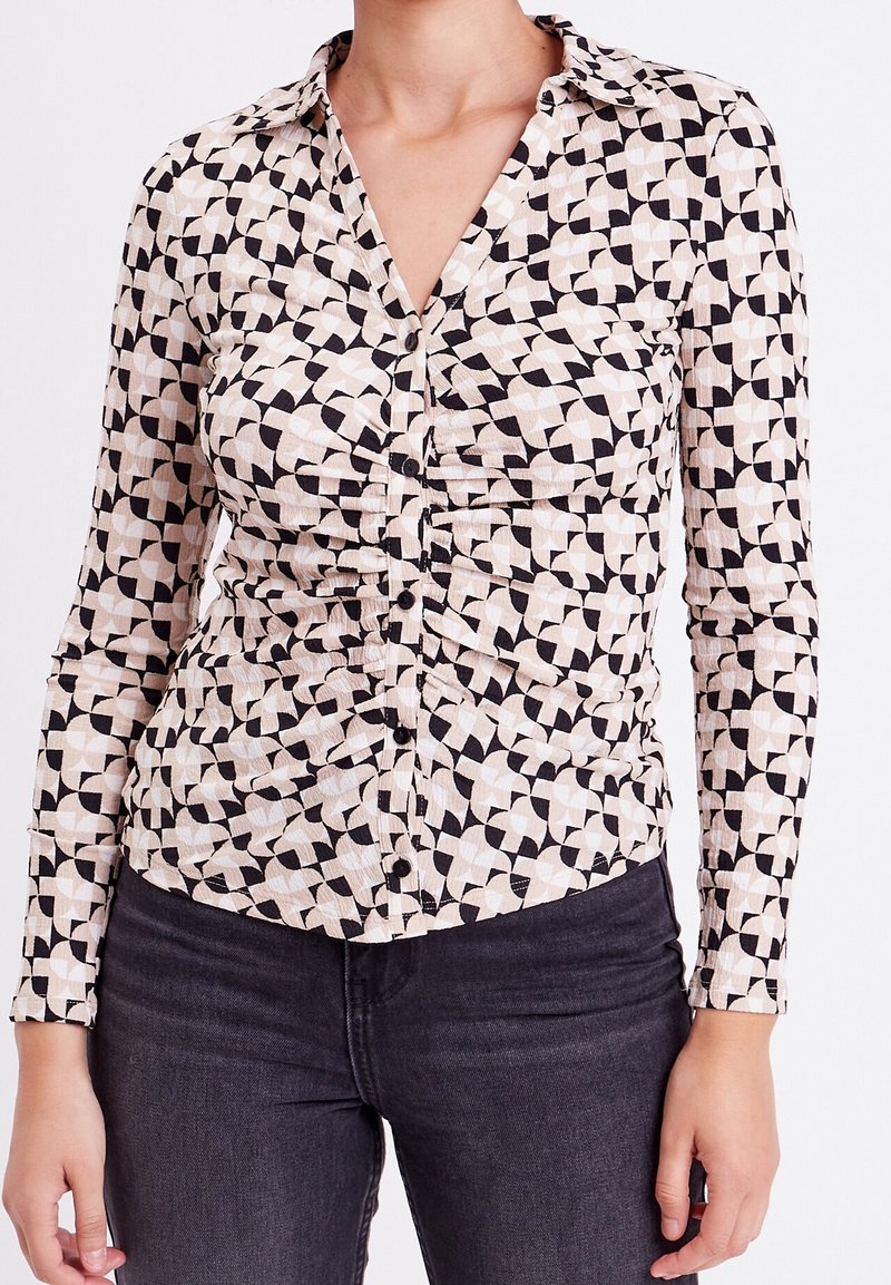 Mujer vistiendo una camisa de botones de manga larga con un patrón geométrico en beige y negro, y jeans oscuros ajustados.