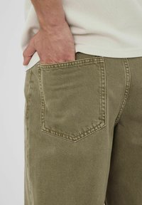 Pantalons olive avec une poche arrière dotée de coutures renforcées. Le matériau est en denim texturé, avec une coupe droite.