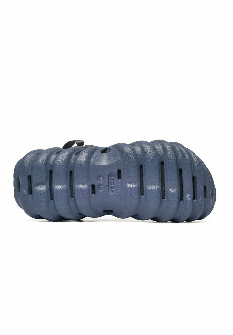 Zapato de goma azul con un diseño segmentado, parte superior texturizada y cortes laterales para ventilación. Presenta una lengüeta para facilitar su uso.