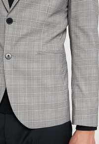 Nära bild av en person som bär en grå och svart rutiga blazer med knappar och en ficka, tillsammans med svarta byxor och en svart skjorta.