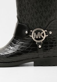 Botte en caoutchouc noire avec un motif crocodile brillant, présentant une tige en tissu sombre texturé et un accent argenté en forme de logo circulaire MK près de la cheville.