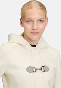 Cremefarvet hættekappe med en tekstureret finish, der har en grå togglesløjfe, synlige sømdetaljer og et hyggeligt, blødt materiale.