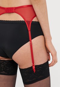 Raudonas tinklelio tipo kabančio diržo diržas su reguliuojamomis petnešėlėmis, su juodos nėriniais išmargintomis kojinėmis ir užsegimu už nugaros su kabliukais.