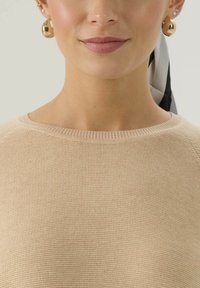 Beżowy sweter z dzianiny z okrągłym dekoltem i widocznymi prążkami. Modelka nosi złote kolczyki i czarno-białą jedwabną chustę.