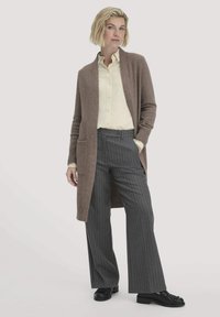 Cardigan lungo marrone con tasche laterali, indossato sopra una camicia gialla chiara. Abbinato a pantaloni grigi a righe sottili e mocassini neri. Tessuto morbido.