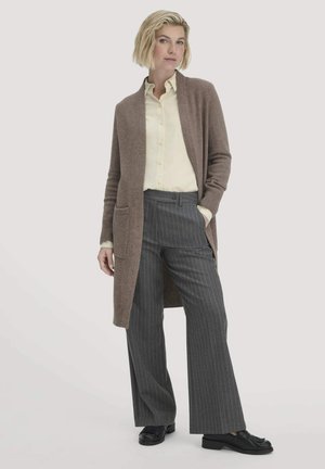 Ein brauner, langer Cardigan mit Seitentaschen, getragen über einem hellgelben Hemd. Kombiniert mit grauen Nadelstreifenhosen und schwarzen Slippern. Glatte Textur.