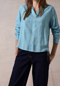 Blusa a maniche lunghe blu chiaro con scollatura a V, caratterizzata da un motivo geometrico in linee nere. Abbinata a pantaloni in denim scuri con dettaglio con zip.