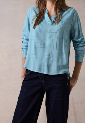 Lyseblå langærmet bluse med V-hals, prydet med geometrisk mønster i sorte linjer. Sammen med mørke denimbukser med lynlåsdetalje.