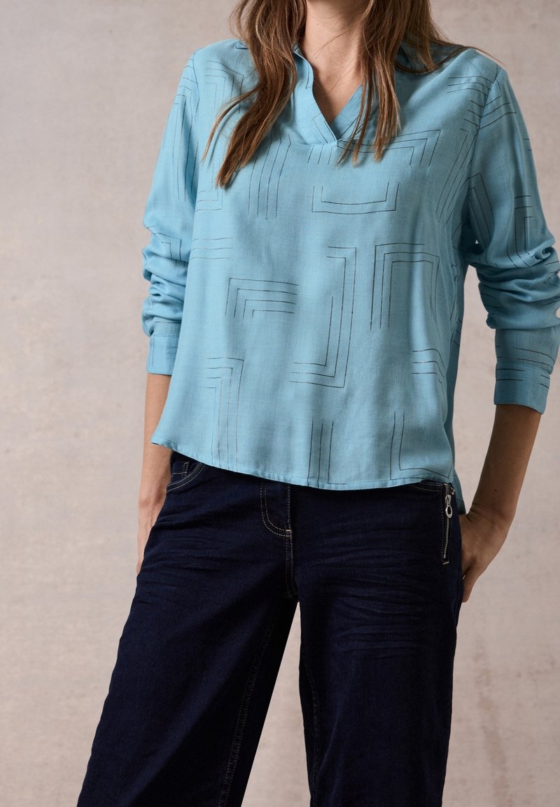 Blusa a maniche lunghe blu chiaro con scollatura a V, caratterizzata da un motivo geometrico in linee nere. Abbinata a pantaloni in denim scuri con dettaglio con zip.