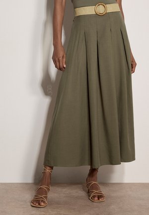 Frau trägt olivgrüne plissierte Hose mit weitem Bein, einen beigen gewebten Gürtel und beige Riemchen-Sandaletten mit Absatz.