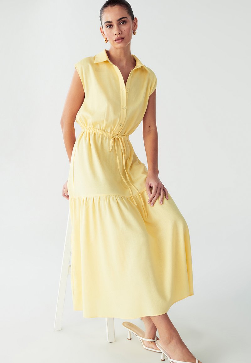 Geltonas, be rankovių, mygtukų užsegimas su raišteliu juosmeniui ir sluoksniuota apačia. Pagamintas iš minkštos medžiagos, pasižymi laisvu siluetu ir apykakle.