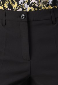 Un pantalon noir doté d'une fermeture à boutons, d'un design à plis plats et de poches latérales. Le tissu semble lisse avec une texture subtile.
