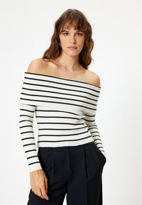 Maglione a spalle scoperte a costine di colore bianco con righe orizzontali nere. Design aderente con maniche lunghe, lavorazione a maglia testurizzata e lunghezza cropped.