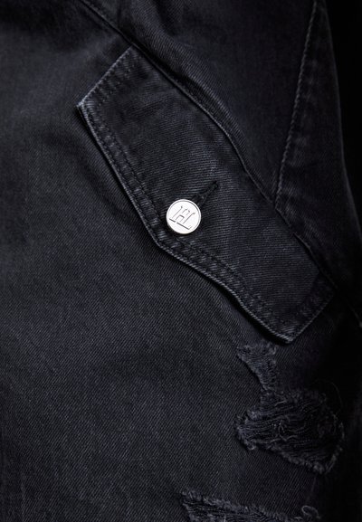 Tecănele de denim negru cu o secțiune uzată, având un nasture metalic argintiu și un buzunar cu clapetă. Textura este netedă, cu un aspect ușor uzat.