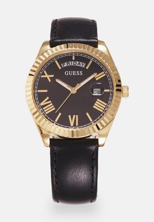 Gold-tone Guess wristwatch with black dial, Roman numerals, day and date display, and black leather strap, showing time 10:10:35 on Friday.
