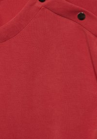 Röd bomulls-tyg med en slät yta, med axeldetalj prydd av svarta tryckknappar och en rundad udd.