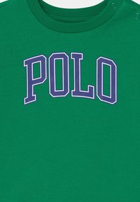 Zelené bavlněné tričko s velkým fialovým nápisem "POLO" na hrudi. Kulatý výstřih s druky na rameni.