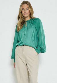 Tee-Bluse in Blaugrün mit eingesammeltem Ausschnitt und langen, puffigen Ärmeln, kombiniert mit beigen Hosen. Der Stoff ist glatt und hat einen leichten Glanz.