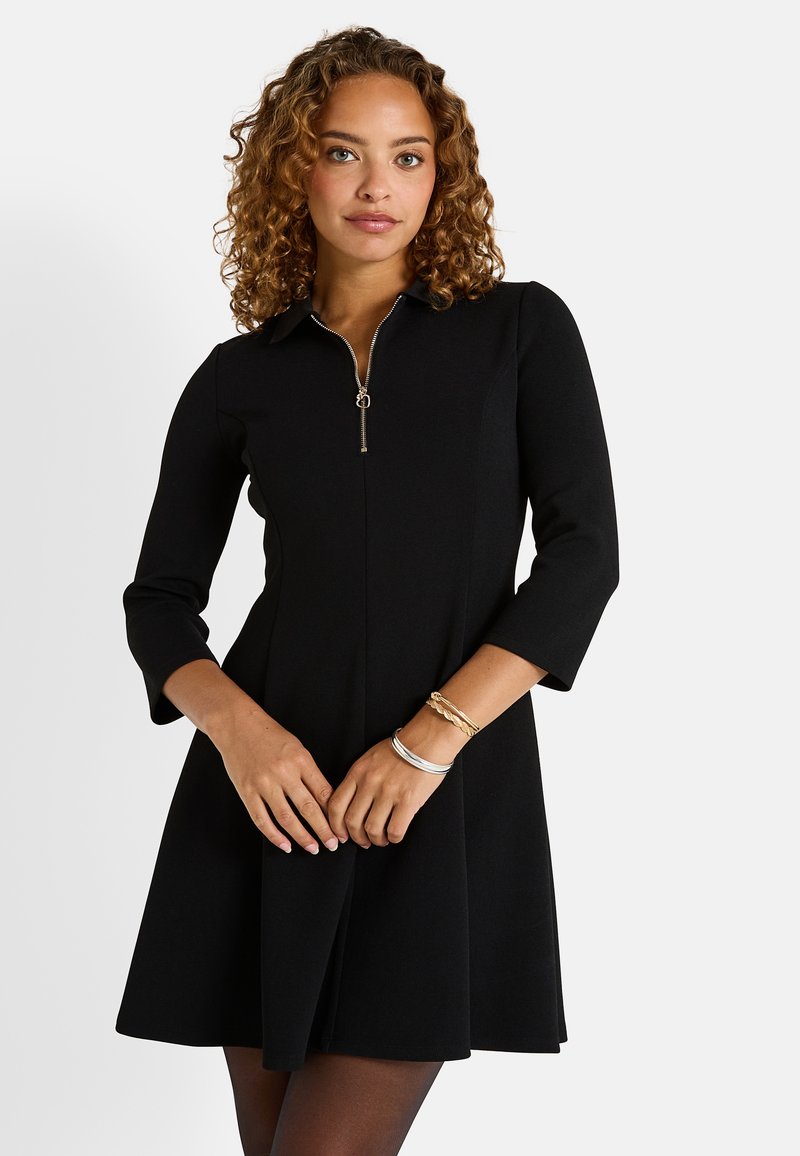 Robe noire avec un col et une fermeture zippée mi-haute, dotée de manches trois-quarts et d'une jupe évasée. Le tissu a l'air lisse et structuré.