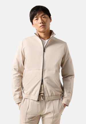 Ung mand iført en beige jakke med lynlås og matchende bukser med hænderne i lommerne, stående mod en ensfarvet baggrund.