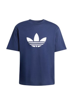 Uomo con dreadlocks che indossa una maglietta Adidas blu navy e pantaloni della tuta Adidas bianchi, in piedi con una mano in tasca.