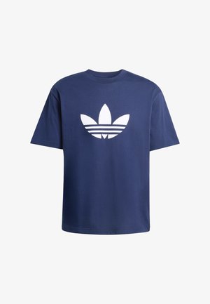 Mężczyzna z dredami, ubrany w granatowy T-shirt Adidas i białe spodnie dresowe Adidas, stoi z jedną ręką w kieszeni.