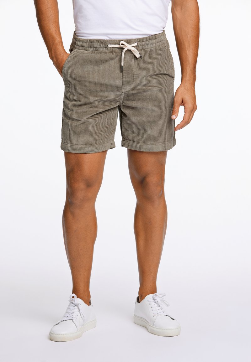 Shorts de pana verde oliva claro con cintura elástica, cierre de cordón, bolsillos laterales y dobladillos enrollados, combinados con zapatillas blancas.