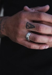 Anello d'argento con un design testurizzato e organico, indossato su una mano che presenta un tatuaggio geometrico. La mano ha un tono di pelle medio.