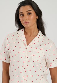 Chemise de pyjama blanche à manches courtes avec une surface texturée et des imprimés coeurs rouges dispersés. Elle comporte une poche poitrine et un col classique.