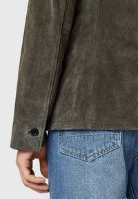 Giacca di camoscio verde oliva con una texture liscia, caratterizzata da un bottone sul polsino e un taglio alla moda, abbinata a jeans di denim blu chiaro.