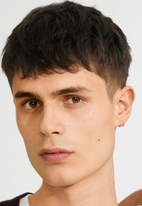 Primo piano di una persona giovane con capelli corti e scuri e occhi nocciola. Indossa piccoli orecchini a cerchio dorati. Pelle liscia con un incarnato chiaro.